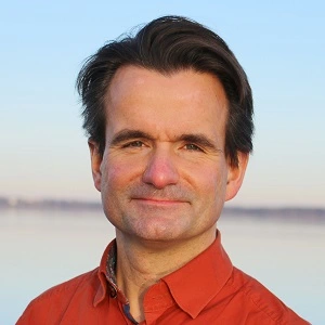 Das Bild zeigt einen Mann mit dunklem, gepflegtem Haar und einem leichten Lächeln. Er trägt ein oranges Hemd, und im Hintergrund ist eine ruhige Gewässerlandschaft bei klarem Himmel zu sehen. Der Mann blickt direkt in die Kamera und vermittelt ein Gefühl von Gelassenheit und Präsenz.