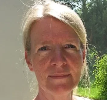 Das Bild zeigt eine Frau mit blondem Haar und ernstem Gesichtsausdruck, die direkt in die Kamera blickt. Sie steht im Freien, wie an der natürlichen, grünen Umgebung im Hintergrund und dem Licht, das auf ihr Gesicht fällt, zu erkennen ist.