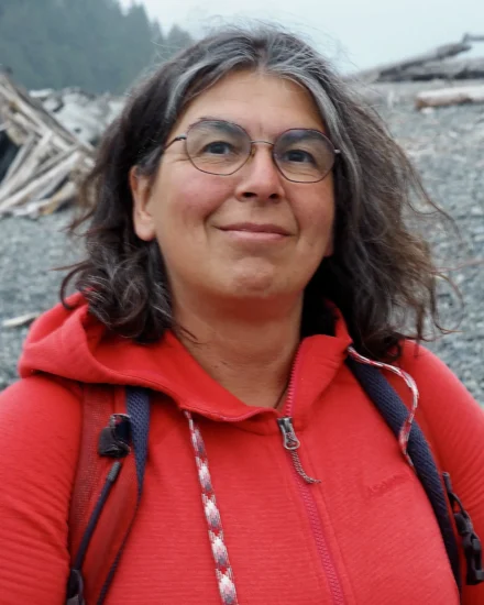 Das Bild zeigt eine Frau mittleren Alters mit schulterlangem, graumeliertem Haar und einer Brille. Sie trägt eine rote Jacke und hat einen Rucksack auf dem Rücken. Ihr freundlicher Gesichtsausdruck und die entspannte Haltung vermitteln eine positive Ausstrahlung. Der Hintergrund zeigt eine steinige Landschaft mit einigen Baumstämmen und Ästen, was darauf hinweist, dass die Frau sich in der Natur aufhält, möglicherweise auf einer Wanderung. Die Umgebung wirkt ruhig und abgeschieden, mit Wald im Hintergrund.