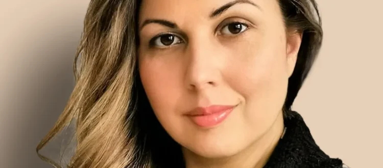 Das Bild zeigt eine Frau mit einem selbstbewussten Blick. Sie hat lange, gewellte Haare mit hellen Strähnen und trägt ein schwarzes Oberteil. Ihr Make-up ist dezent, betont aber ihre Gesichtszüge, insbesondere ihre Augen. Der Hintergrund ist neutral und lässt die Frau klar hervortreten. Sie wirkt professionell und zugleich modern.